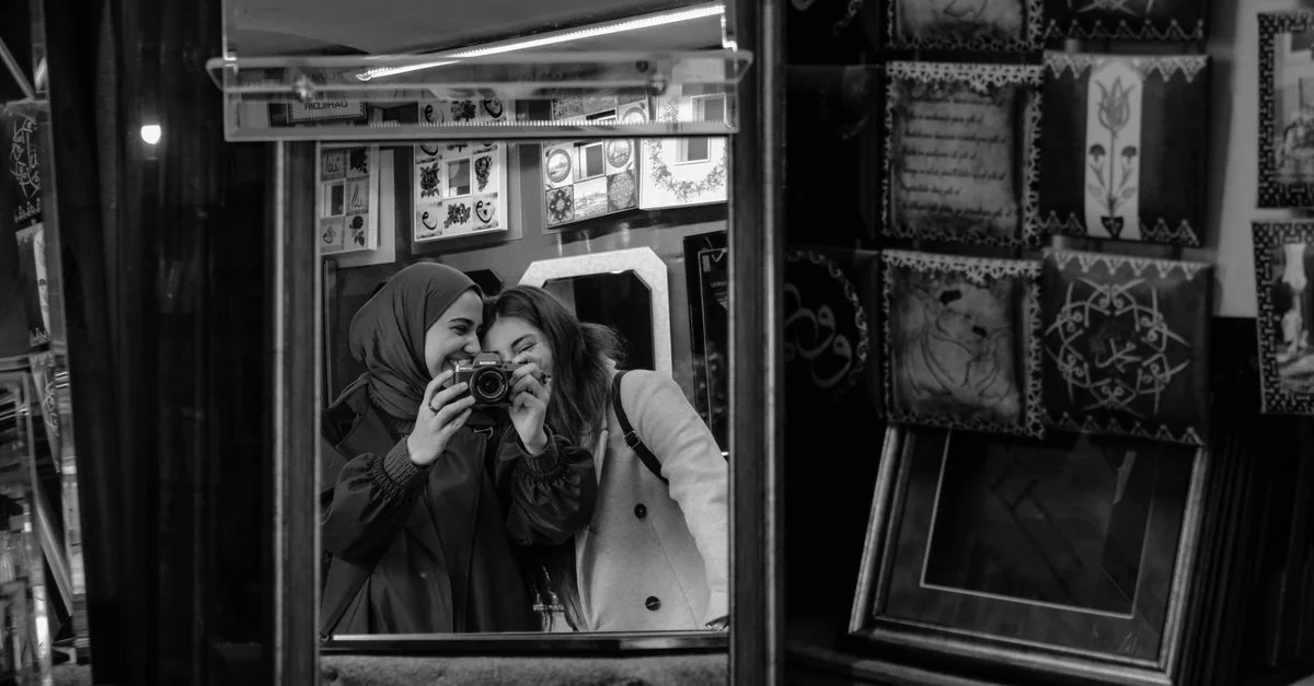 Dos mujeres sonriendo en un espejo, capturando un momento con una cámara en una tienda llena de arte.