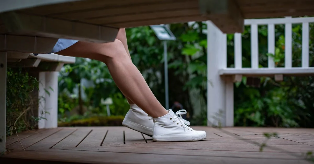 Las piernas de una mujer con zapatillas blancas descansando en un banco de madera en un jardín verde y exuberante.