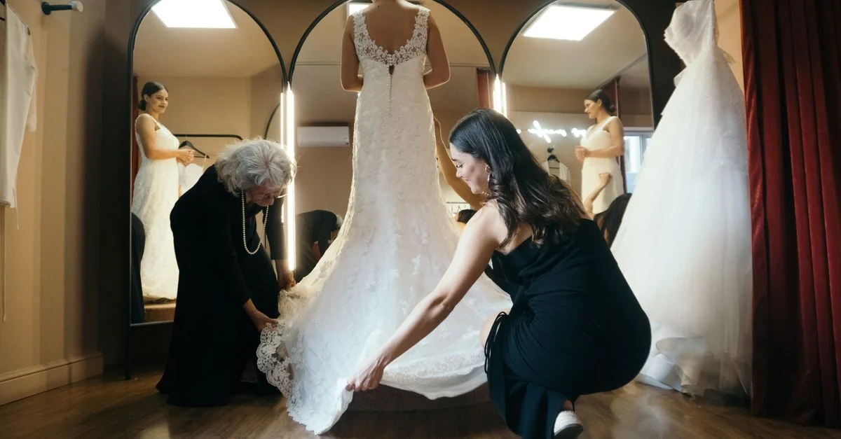 Una novia se encuentra en un vestidor mientras dos mujeres ayudan con los ajustes de su vestido de novia.