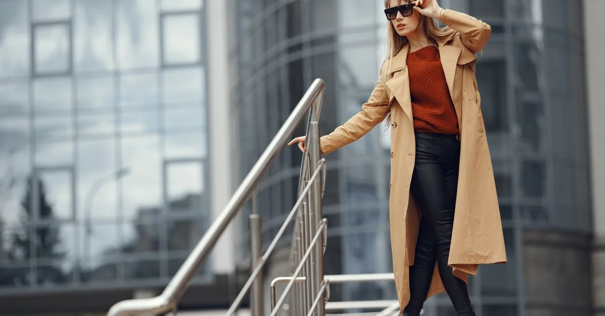 Mujer elegante con abrigo de trench ajustando gafas de sol en escaleras de la ciudad con fondo de edificio de oficinas.