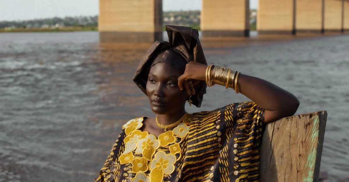 Una mujer con atuendo tradicional africano, sentada junto a un río con un puente al fondo, irradiando gracia.