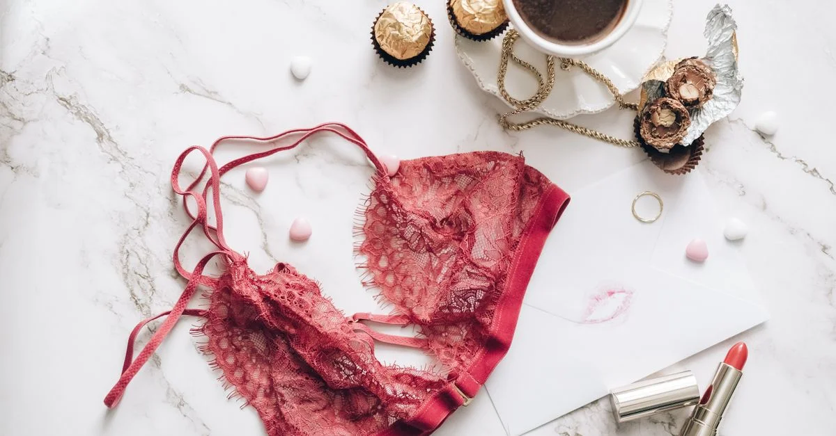Flat lay elegante con lencería de encaje rojo, café, chocolates y pintalabios sobre una superficie de mármol.